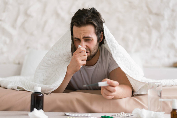 Despaired sad young caucasian sick male lies on bed under blanket, suffers from runny, blows his nose in napkin in white bedroom interior. Illness, flu and cold, bad feeling and treatment at home - Foto, imagen