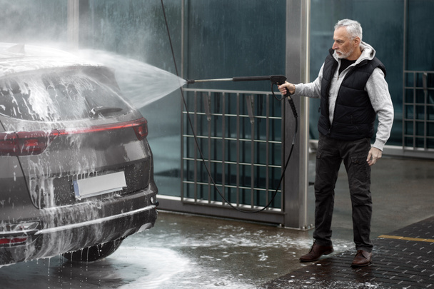 Full-length portrait of elderly Caucasian white-bearded man, washing off the detergent foam soap suds from his car with water from pressurized hose, at a self-service car wash station. Car maintenance - Photo, Image