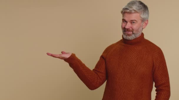 Handsome middle-aged mature man showing thumbs up and pointing empty place, advertising area for commercial text, copy space for goods promotion. Senior guy. Studio shot indoors on beige background - Footage, Video