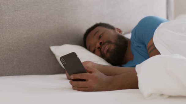 Internet addiction concept. Young indifferent african american guy web surfing on smartphone, cant fall asleep, lying in bed at home, slow motion, empty space - Footage, Video
