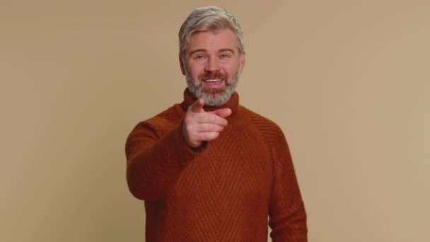 I choose you. Middle-aged mature man pointing to camera and looking with playful happy expression, making choice, showing direction. Senior smiling old guy isolated alone on beige studio background - Filmati, video
