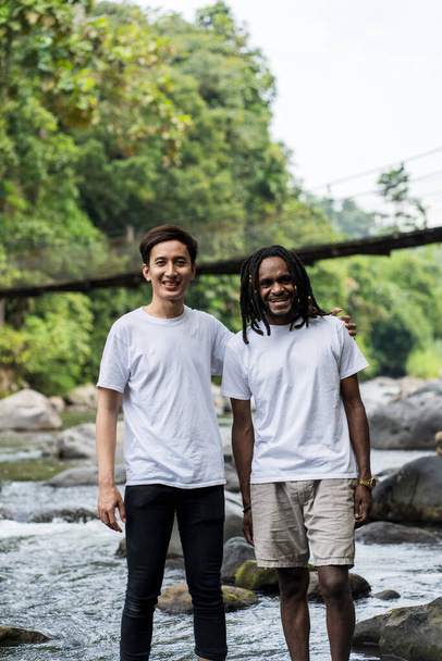 Азіатський і африканський чоловік з білою порожньою сорочкою, що стоїть, посміхаючись з річкою як тлом у природі
 - Фото, зображення