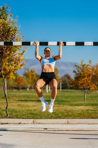 Strong fit girl doing pull ups while holding the goal's crossbar. Working out in the park - Foto, imagen