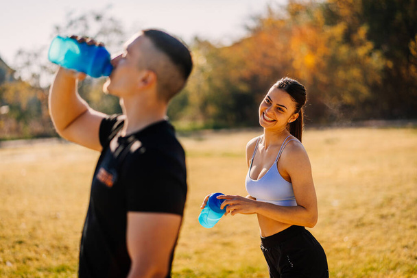 Een mooi fit meisje lacht en kijkt naar haar vriend terwijl hij water drinkt - Foto, afbeelding