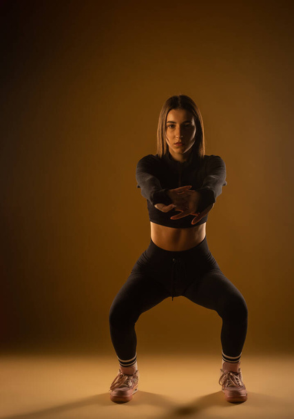 Cute young girl in black sportswear doing squats in studio - Foto, imagen
