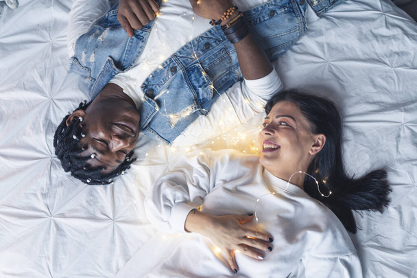 Fröhliches gemischtrassiges Paar, das mit weißen Laken auf seinem Bett liegt. Kaukasische junge erwachsene Frau, die ihrem geliebten afroamerikanischen Partner in die Augen schaut. Paar mit Weihnachtsbeleuchtung. Hohe Qualität - Foto, Bild
