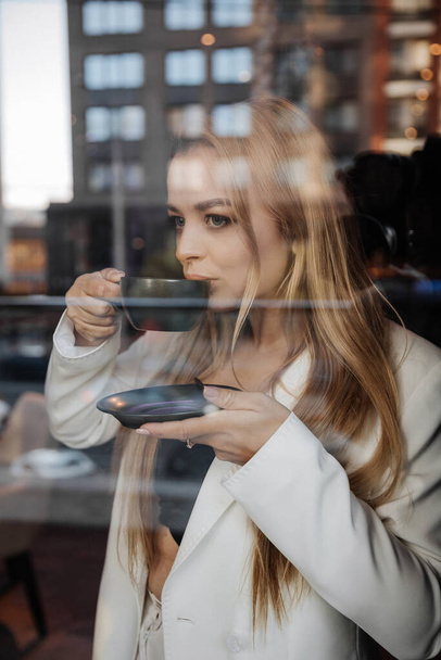 Blond stijlvolle vrouw bij oversized witte jas drinken aroma koffie en ontspannen op de moderne stad achtergrond - Foto, afbeelding