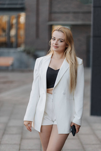 Blond stylish woman at oversized white jacket and phone on hand posing at the railing and steps on the city background - Foto, imagen