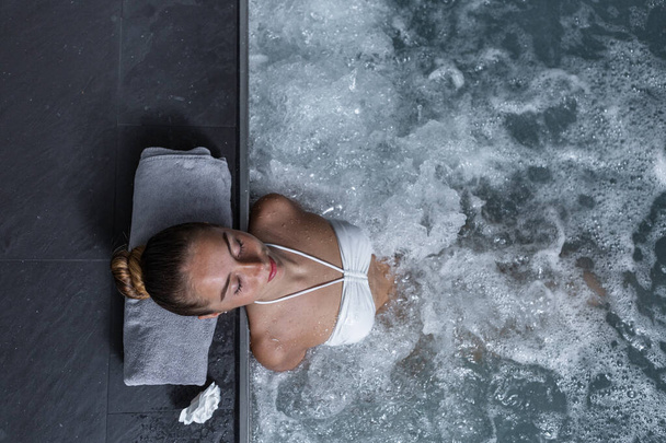 Top view of young woman with closed eyes leaning on pool border with towel and enjoying hydrotherapy in clean pool with splashing water - Photo, image