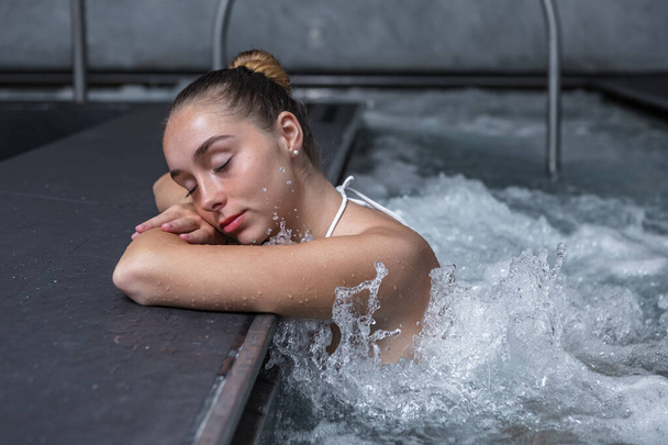 Расслабленная молодая женщина закрывает глаза и держит голову на скрещенных руках, расслабляясь в брызги воды массажного бассейна в выходные дни на курорте - Фото, изображение