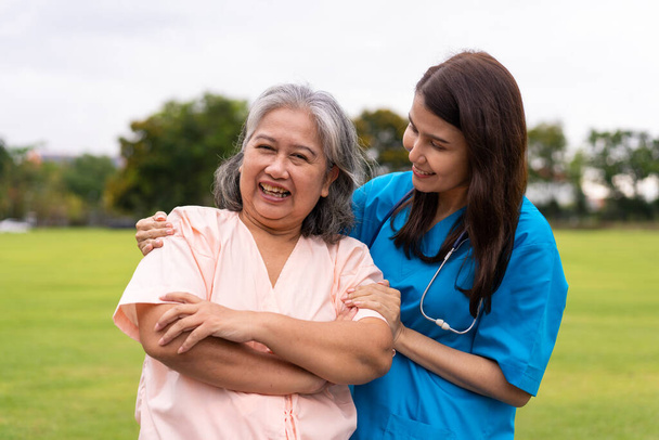 Ασιάτης προσεκτικός φροντιστής ή νοσηλευτής κρατά το χέρι του ασθενούς και ενθαρρύνει τον ασθενή σε έναν κήπο. Έννοια της ευτυχούς συνταξιοδότησης με φροντίδα από έναν φροντιστή και Ταμιευτήριο και την ανώτερη ασφάλιση υγείας. - Φωτογραφία, εικόνα