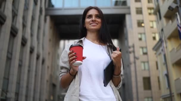 Успішна молода красива молода жінка з планшетом і чашкою кави ходить між бізнес-будівлями і посміхається з радістю. Приваблива ділова дівчина. Кар'єра людей
. - Кадри, відео