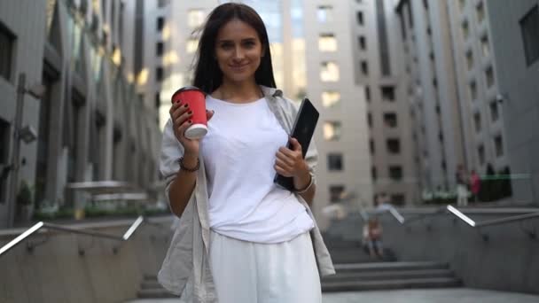 Sikeres fiatal, gyönyörű fiatal nő táblagéppel és kávéscsészével sétál az üzleti épületek között, és örömmel mosolyog. Vonzó üzletasszony. Karrier emberek. - Felvétel, videó