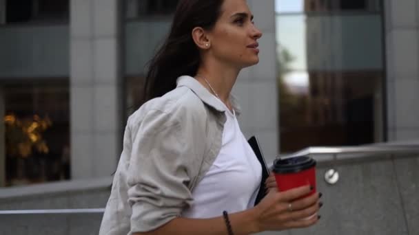 Sikeres fiatal, gyönyörű fiatal nő táblagéppel és kávéscsészével sétál az üzleti épületek között, és örömmel mosolyog. Vonzó üzletasszony. Karrier emberek. - Felvétel, videó