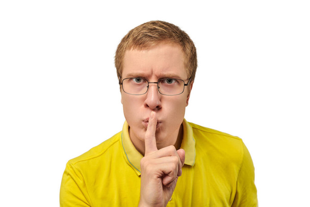 Funny young man in yellow T-shirt asking to be quiet, silence gesture isolated on white background. Young man in glasses saying Shhh, keep quiet, please and making silence gesture, request for silence - Photo, image