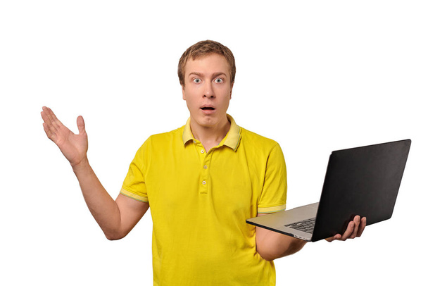 Здивований хлопець тримає ноутбук в руці ізольовано на білому тлі. Смішний шокований молодий чоловік у жовтій футболці тримає блокнот в руці. Смішний хлопець з спантеличеним виглядом через продажі та знижки
 - Фото, зображення