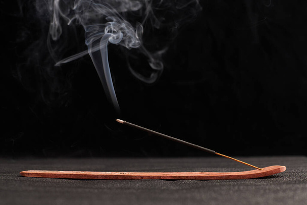 Smoke curls of incense stick in wooden holder for relaxation and meditation exercises, black background. Aromatherapy session with burning aroma stick, pleasant aroma and incense - Photo, Image