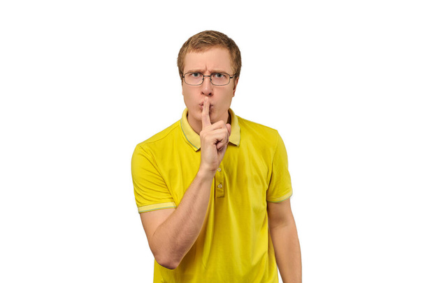Забавный молодой человек в желтой футболке, просящий тишины, жеста молчания изолированы на белом фоне. Молодой человек в очках говорит: Тихо, пожалуйста, и делая жест молчания, просьба о молчании - Фото, изображение