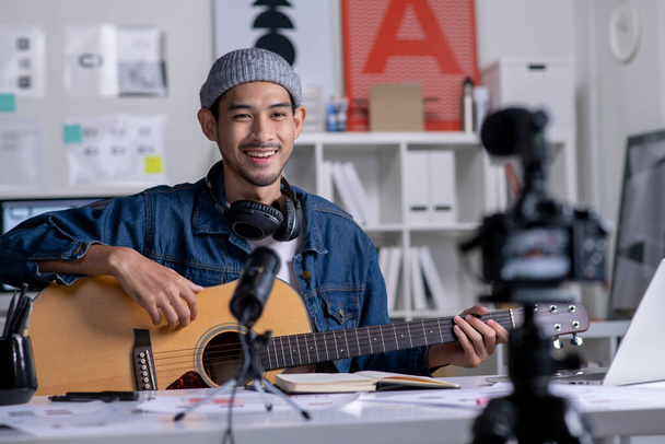 Young Asian male graphic designer blogger influencer playing guitar talking looking at camera while shooting education tutorial vlog training filming video course for social media at studio. - Zdjęcie, obraz