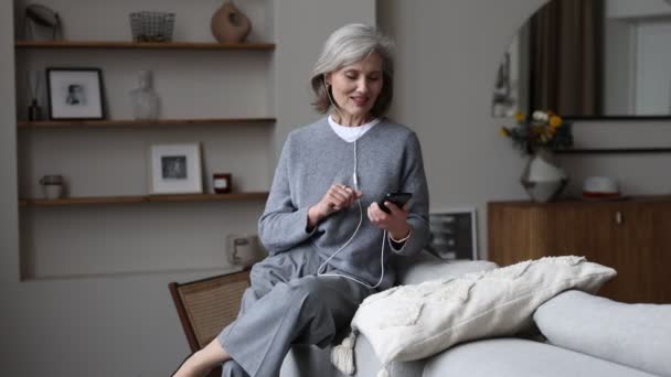 volwassen stijlvolle vrouw op communiceert via videogesprek met behulp van telefoon in hoofdtelefoon terwijl thuis zitten. - Video