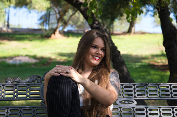 Hermosa joven con el pelo rubio y ojos azules se sienta en el banco del parque y posa para la foto como una modelo. Ella es feliz haciendo diferentes posturas y expresiones corporales. - Foto, Imagen