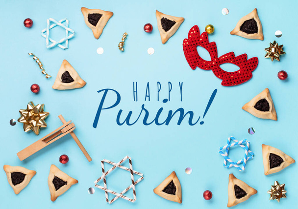 Purim celebration jewish carnival holiday concept. Tasty hamantaschen cookies, red carnival mask, noisemaker, sweet candies and party decor on blue background. Top view, flat lay, copy space. - Photo, Image