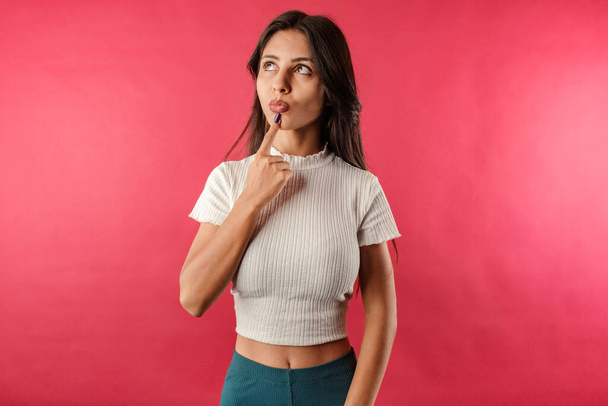 Portrait of young brown-haired woman wearing ribbed crop isolated over red background undecided, purses lips and touches with index finger. Looks at the empty copy space and tries to decide. - Foto, Imagen