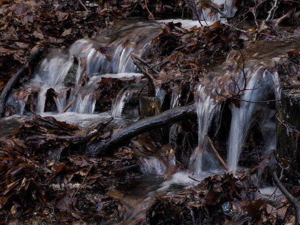 Ένα όμορφο μικρό ρέον ποτάμι λόγω του χιονιού λιώνει σχηματίζοντας ένα καθαρό καταρράκτη με φρέσκο δροσερό νερό που περιβάλλεται από βλάστηση σε ένα άγριο πέτρινο βουνό - Φωτογραφία, εικόνα