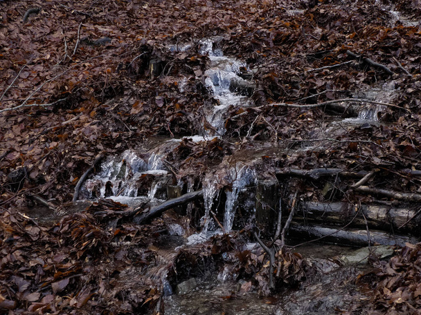 Ένα όμορφο μικρό ρέον ποτάμι λόγω του χιονιού λιώνει σχηματίζοντας ένα καθαρό καταρράκτη με φρέσκο δροσερό νερό που περιβάλλεται από βλάστηση σε ένα άγριο πέτρινο βουνό - Φωτογραφία, εικόνα