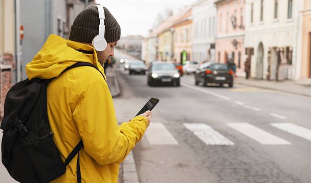 Man in yellow jacket and headphones standing next to road in city using his smart phone while waiting taxi. - Photo, Image