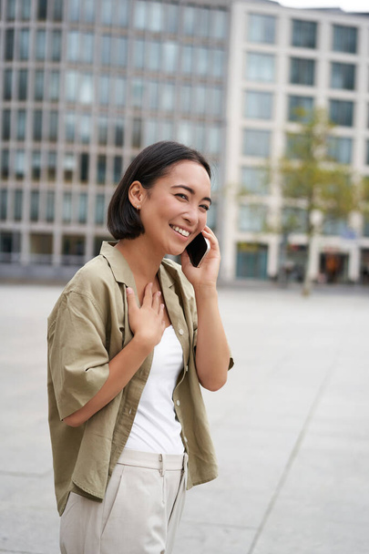 Colpo verticale di sorridente ragazza asiatica elegante, avendo una conversazione telefonica e camminare per strada. Giovane donna che parla sul cellulare. - Foto, immagini