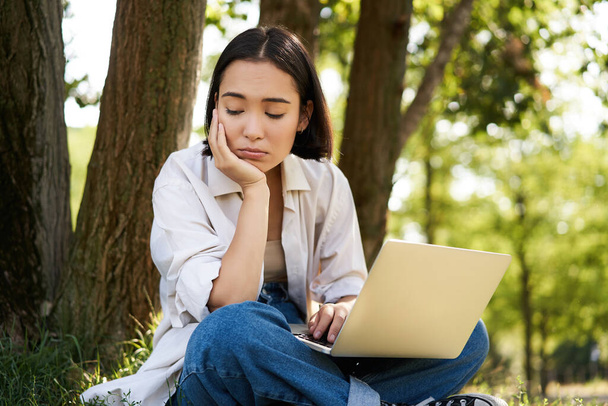 Porträt einer asiatischen Frau, die auf Gras neben Baum sitzt, Laptop benutzt, arbeitet, an sonnigen Sommertagen ferngesteuert Hausaufgaben macht. - Foto, Bild