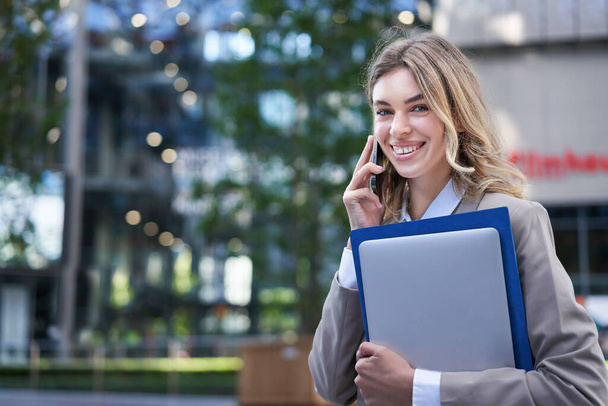 Усміхнена професійна бізнес-леді розмовляє на мобільному телефоні, тримає ноутбук і робочі папки в руці, має діловий дзвінок
. - Фото, зображення