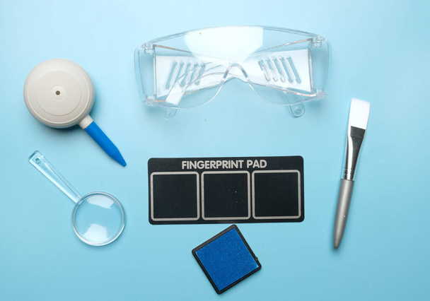 Flatlay picture of toy fingerprint pad, magnifying glass, puffer, brush, safety glass and inkpad on blue background. - Foto, Bild