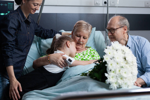 Little granddaughter giving flowers bouquet to sick grandmother. Old female patient receiving floral present from relatives on hospital visit. Girl hugging sick old woman at medical clinic bedroom. - Photo, Image