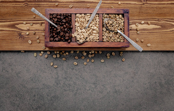 Grains de café verts et bruns non torréfiés et foncés torréfiés dans une boîte en bois avec cuillères sur fond de béton foncé. - Photo, image