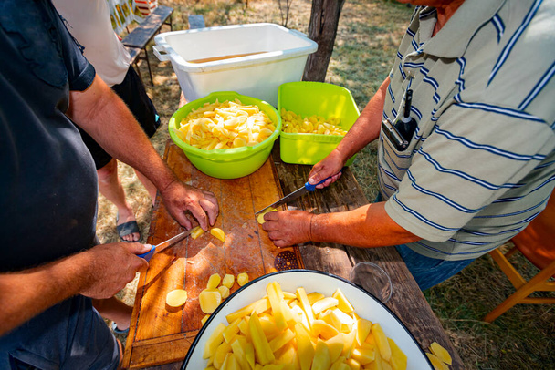 Teamwork of two farmer cookers cut potatoes using knives into slices on the wooden table prepare it for cooking at the outdoor. Bowls are full of cut vegetable  - Photo, image