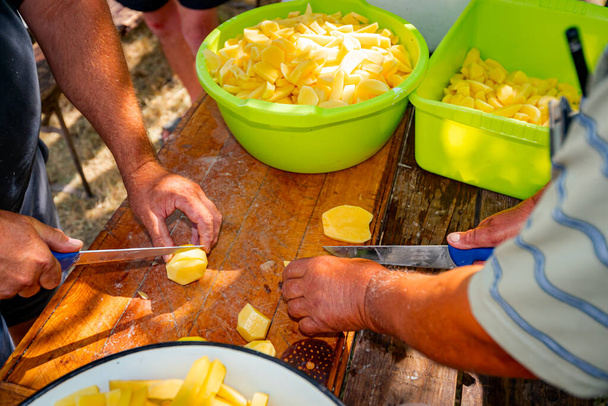 Teamwork of two farmer cookers cut potatoes using knives into slices on the wooden table prepare it for cooking at the outdoor. Bowls are full of cut vegetable  - Foto, immagini