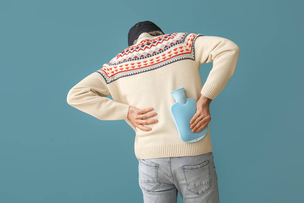 Young man warming his lower back with hot water bottle on color background - Fotoğraf, Görsel