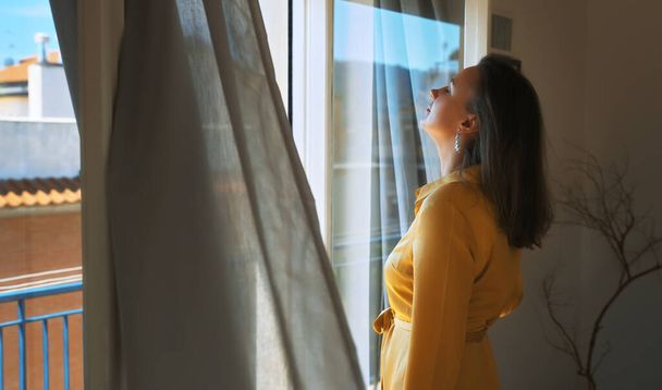 Женщина наслаждается теплым летним бризом, стоя перед окном. - Фото, изображение
