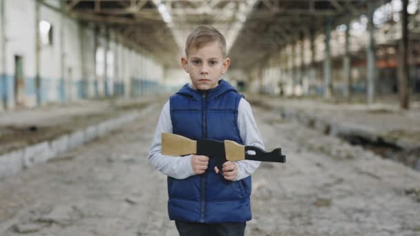 Child holds a toy gun standing against a background of devastation looking at camera. Portrait. Boy games. War. Boy playing in war - Séquence, vidéo