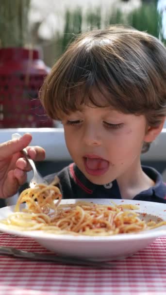 Het kind zit aan tafel spaghetti te eten. Moeder hand voedt pasta aan zoon in restaurant. Noedels met rode saus op bord in verticale video - Video