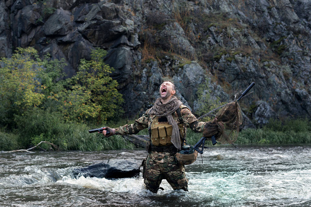 Lone mercenary soldier stands knee-deep in river and screaming, arms outstretched with weapons - he lost all his friends. Despair and helplessness. Concept of modern military and sabotage operations. - Photo, image