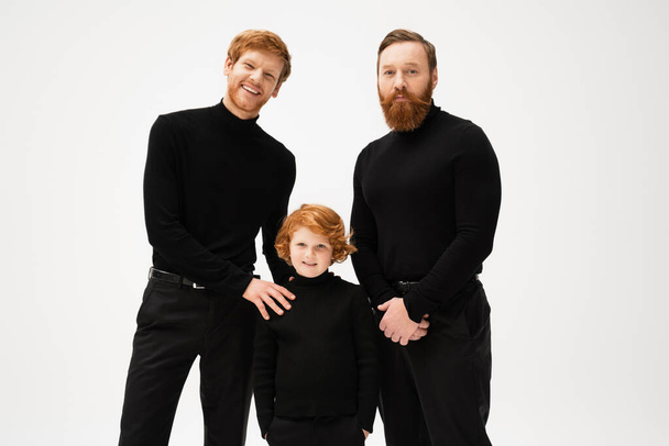 redhead boy standing with hands in pockets near happy dad and grandpa while smiling at camera isolated on grey - Foto, immagini