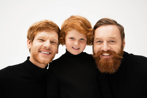 cheerful red haired boy with bearded grandfather and dad in black turtlenecks smiling at camera isolated on grey - Zdjęcie, obraz