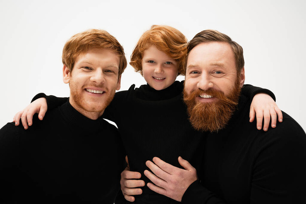 smiling redhead kid hugging bearded grandpa and dad wearing black turtlenecks isolated on grey - Photo, image