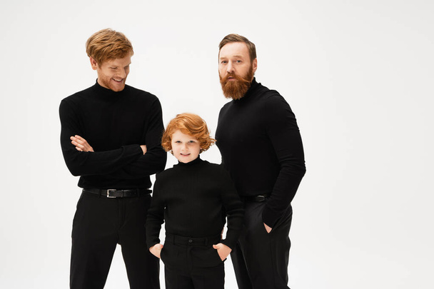 bearded man looking at camera near redhead son and grandson posing in black clothes isolated on grey - Fotó, kép