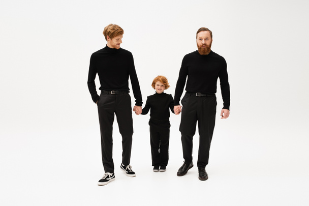 full length of bearded men in black turtlenecks and trousers holding hands with happy red haired kid on light grey background - Foto, Imagem