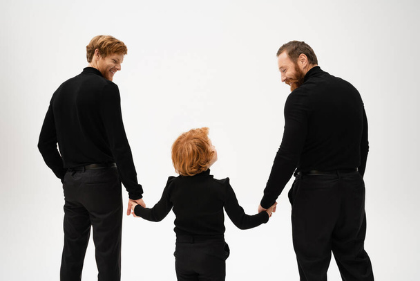 Вид сзади на бородатых мужчин и рыжего волосатого мальчика, держащегося за руки и смотрящего друг на друга изолированно на сером фоне - Фото, изображение