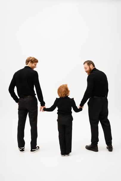 full length of bearded men in black clothes smiling at redhead boy while holding hands on light grey background - Fotoğraf, Görsel